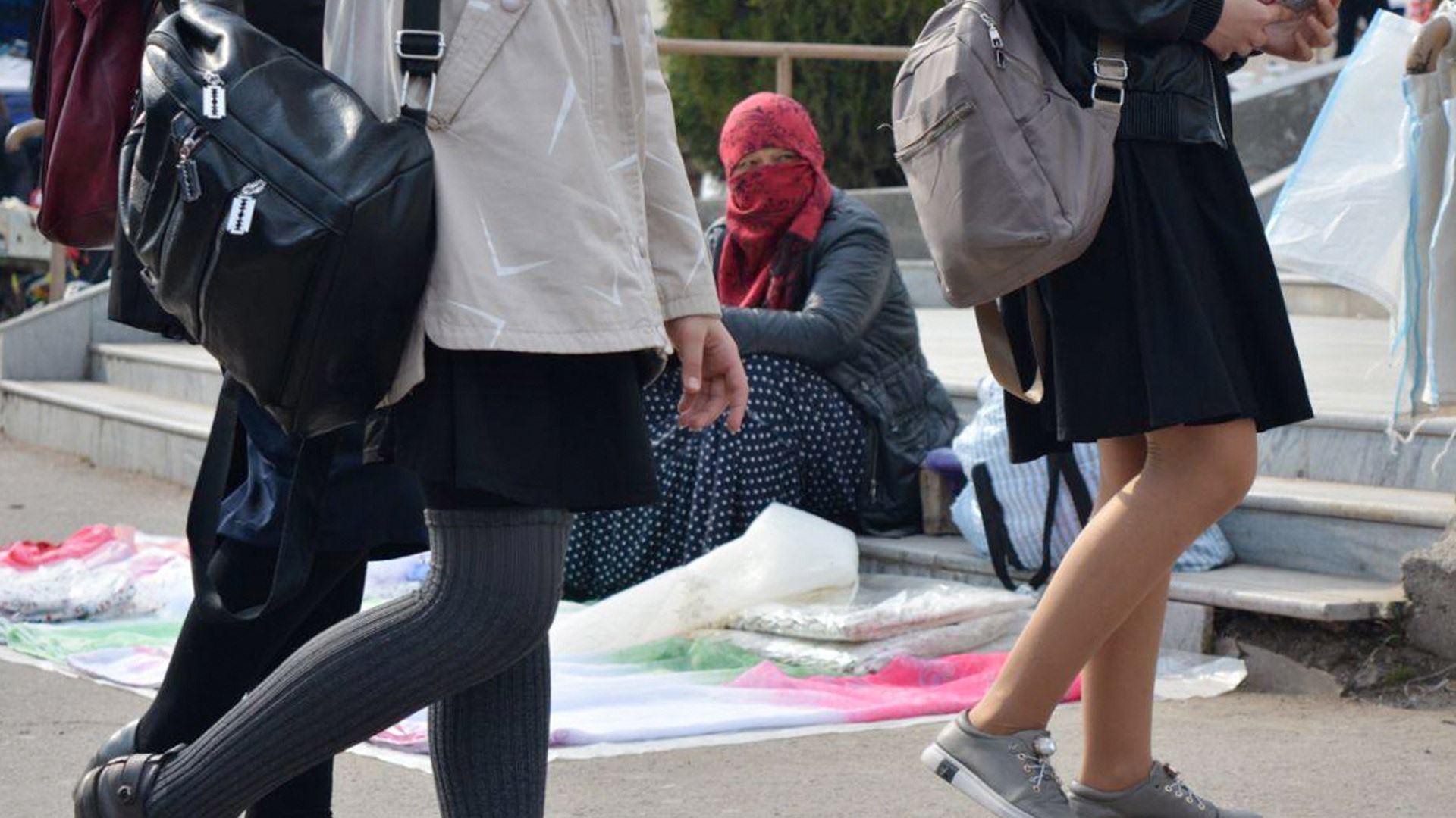 Young people walking past a hijab wearing woman sat on the ground
